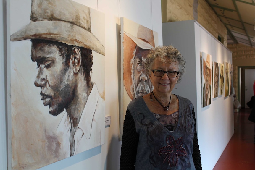 Artist Betty Davies stands in front of a row of paintings of aboriginal stockmen and their hats