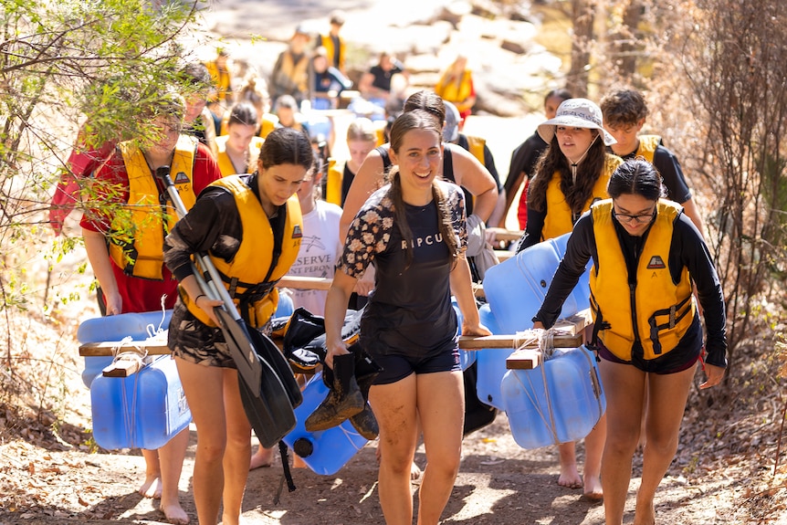 Students wearing life jackets carrying home-made raft and oars up a slight hill.