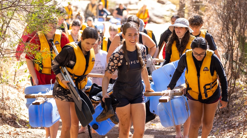 Students wearing life jackets carrying home-made raft and oars up a slight hill.