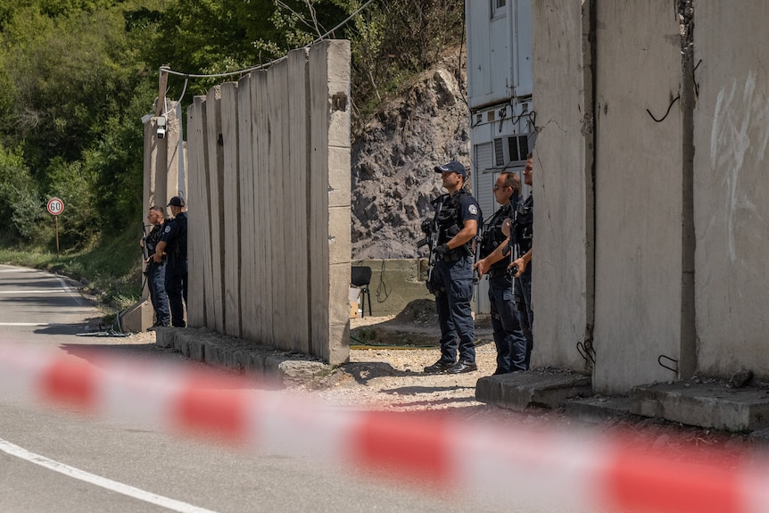 Men in dark clothing hold assault rifles as they stand guard near a wall.