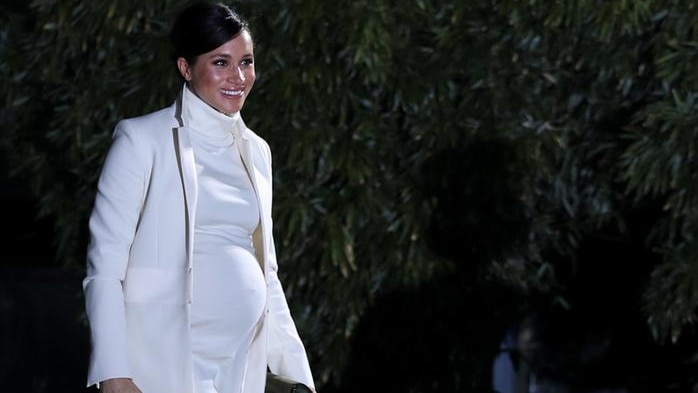 Meghan wears a white turtleneck dress and white coat. She is smiling as she walks with a black purse in hand and hair tied back
