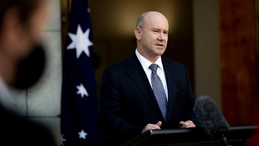Secretary of Defence Greg Moriarty during a press conference. He is wearing a suit and standing at a lectern.