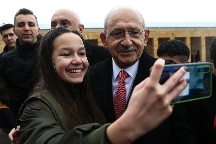 A bald man wearing glasses smiles as he poses with a girl holding a phone to take a selfie.