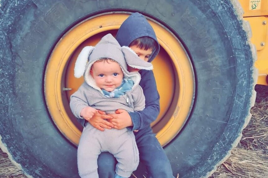A five-year-old boy in black clothes sits in a tractor wheel while holding his five-month-old brother in a rabbit onesie.