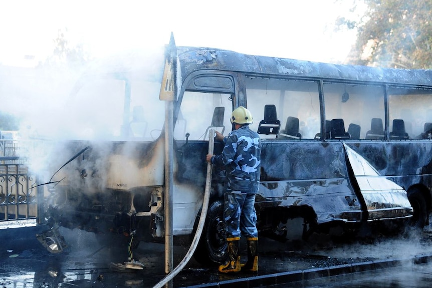 A man in blue and white military uniform hoses down a smoking, charred bus 