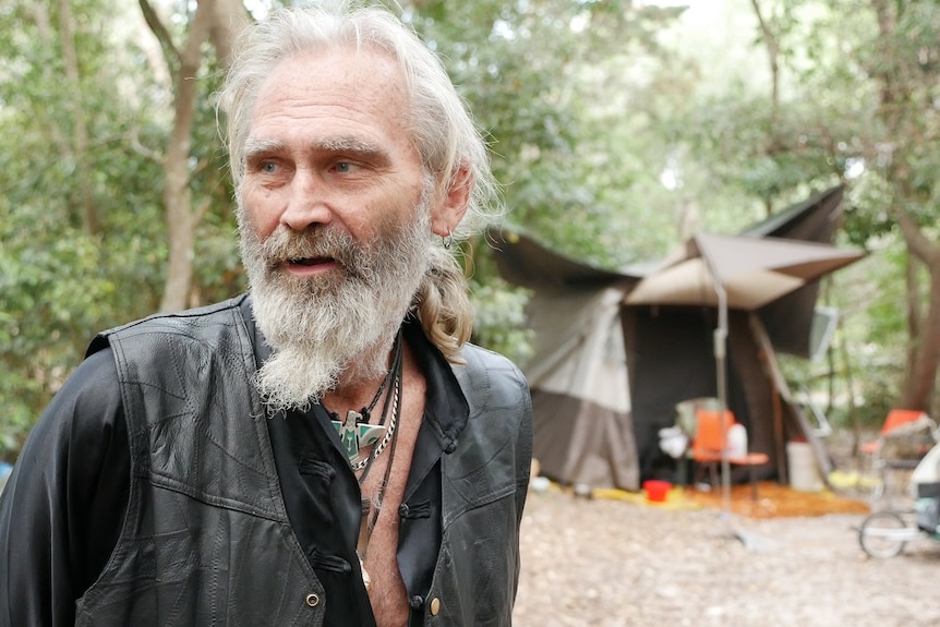 A grey-haired, bearded man dressed in a leather vest looks off-camera at a bush campsite.
