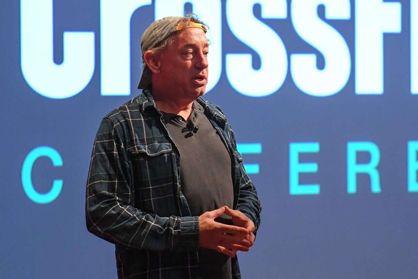 Greg Glassman stands in front of a screen that reads "crossfit conference" wearing a backwards cap and a flannelette shirt.