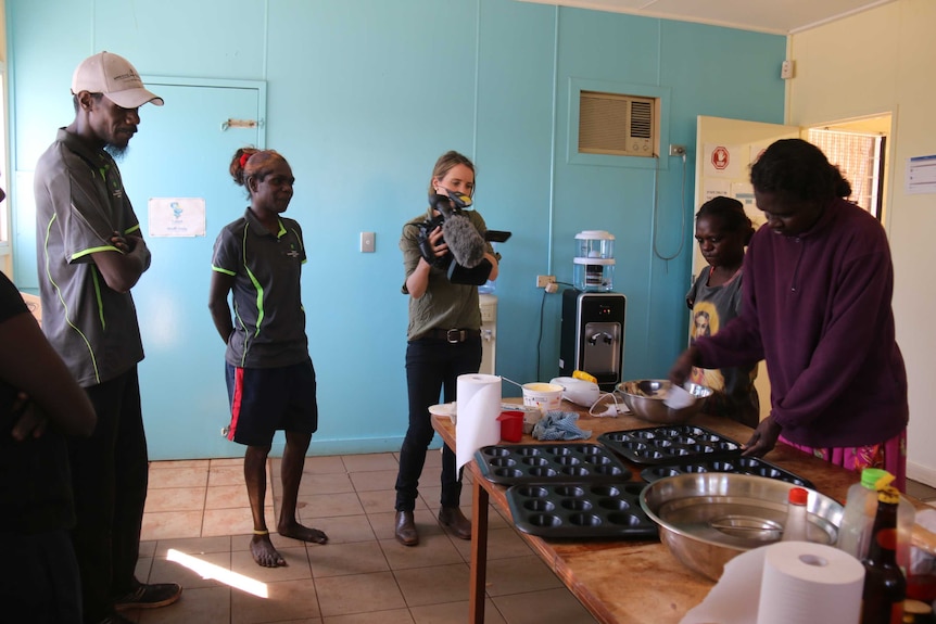 A journalist films women at a table who are making cupcakes.
