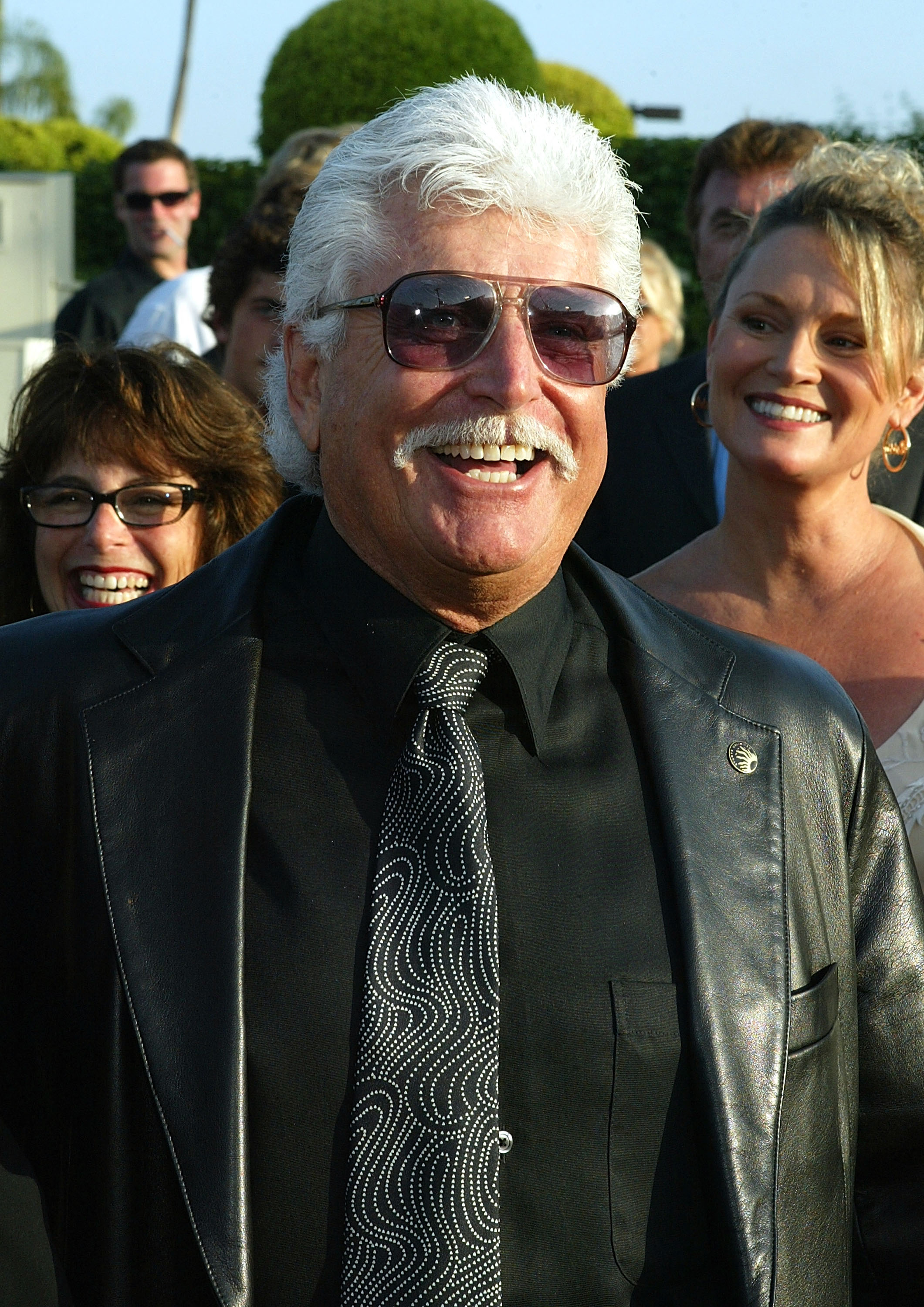 A silver-haired man wearing aviator shades smiles broadly