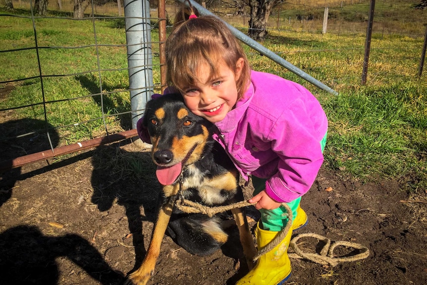 A young girl leaning down, smiling and hugging her kelpie dog tightly