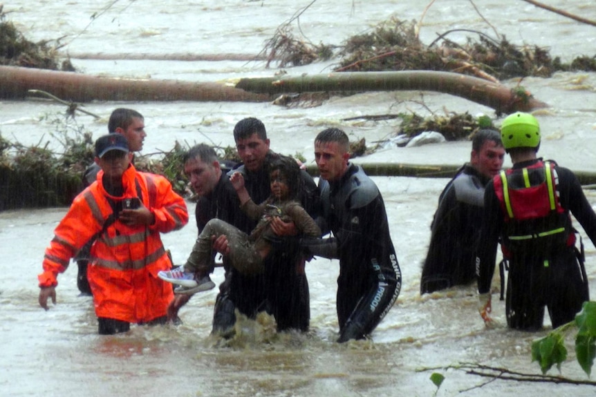 Hombres con trajes de neopreno cargan a una niña a través del agua de una inundación que les llega hasta los muslos. 