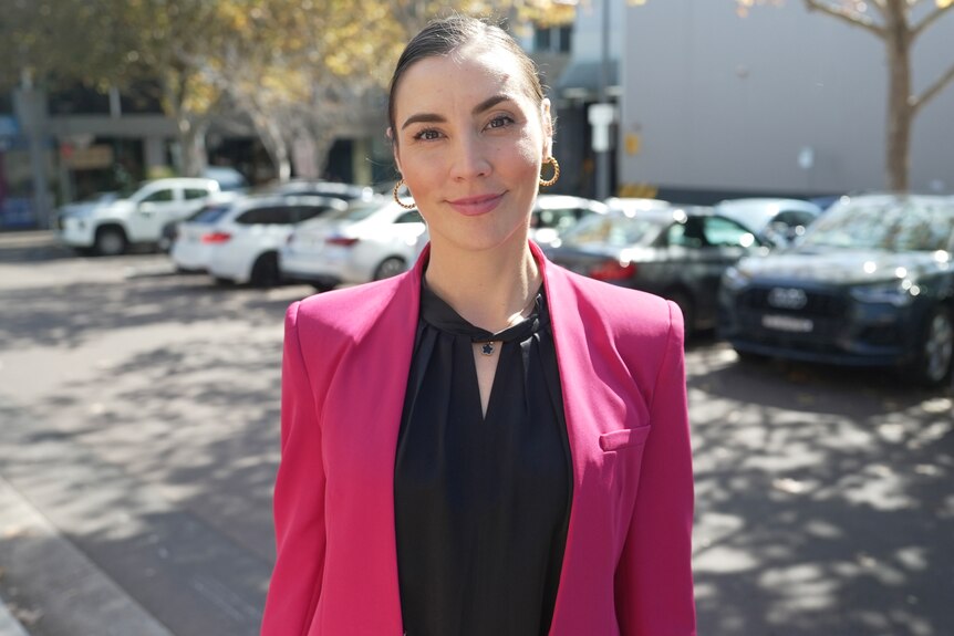 A woman with slicked back brown hair wearing a black shirt and pink blazer smiles while standing in a car park.