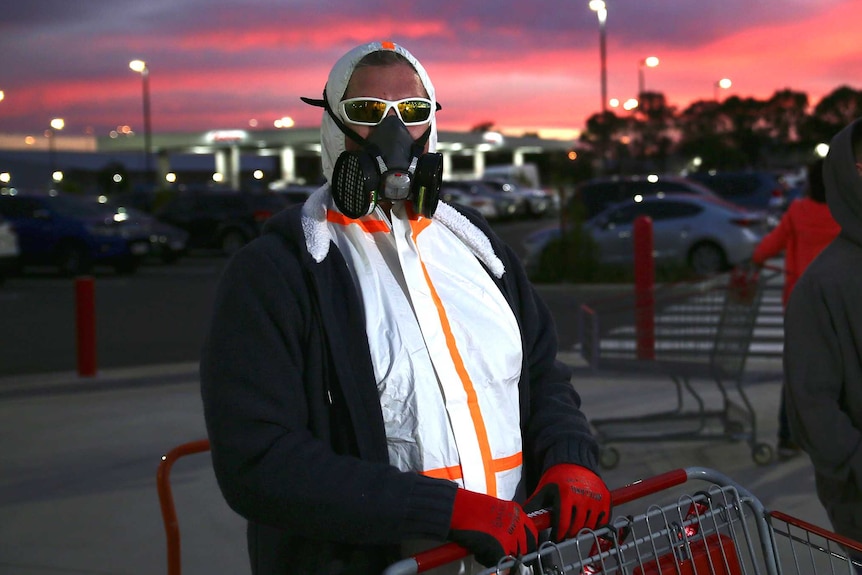A man in a mask and protective suit stands next to a shopping trolley as the sun rises behind him.