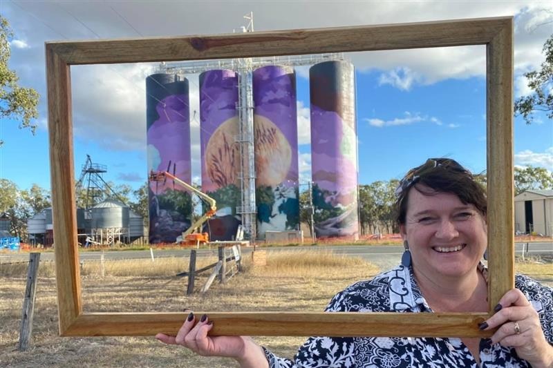 A woman stands smiling, holding a wooden frame around her face and four colourful silos in the background.