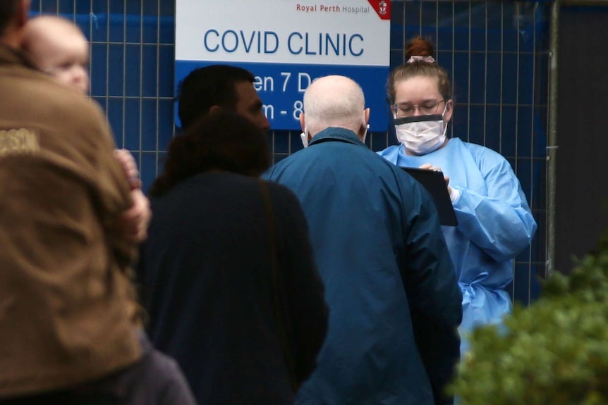 People queue before a woman in a mask and medical scrubs holding a tablet. A sign says COVID Clinic.