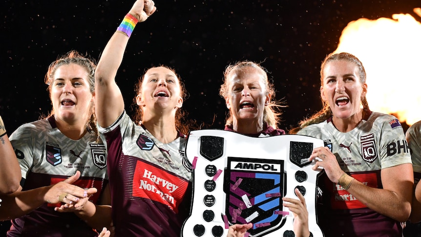 Three Queensland Maroons players and their coach stand and celebrate with the women's State of Origin shield