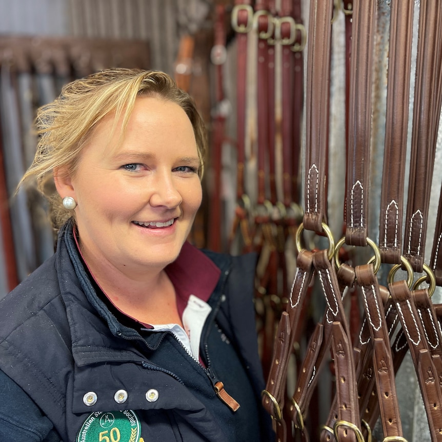 A woman standing a shop next to a rack of leather horse equiptment.