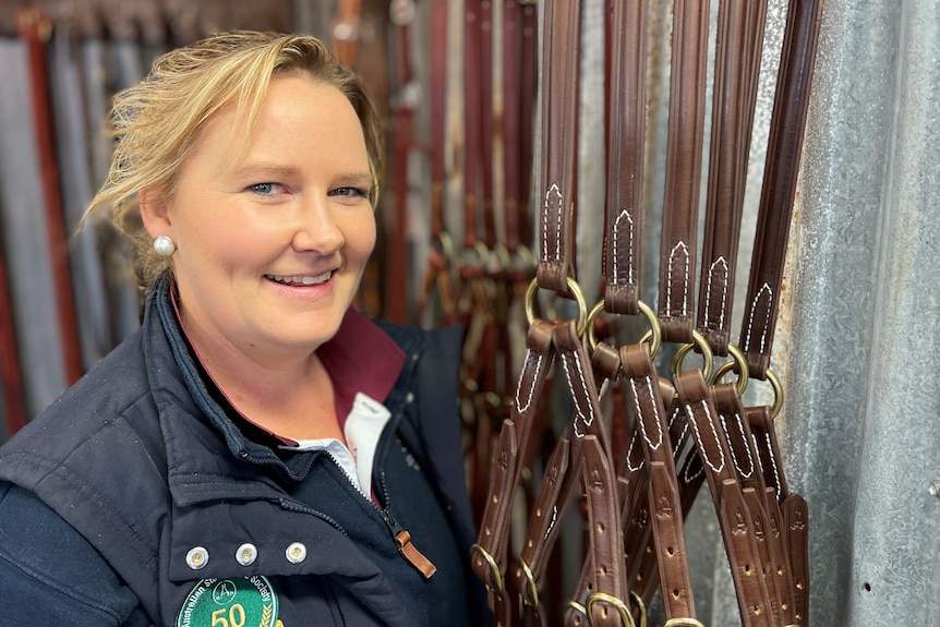 A woman standing a shop next to a rack of leather horse equiptment.