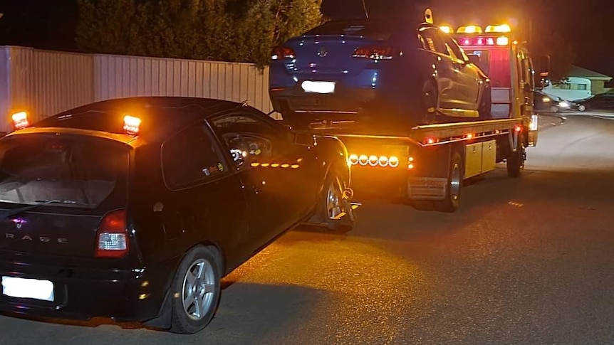 A blue sedan on the back of a flat-bed tow truck with another black hatchback being towed behind