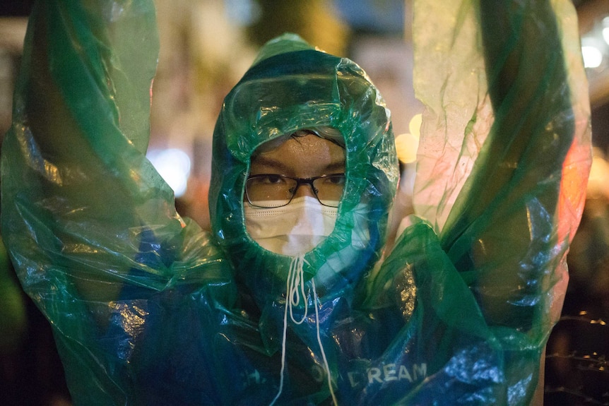 A protester in glasses and facemask wears a raincoat and lifts their arms.