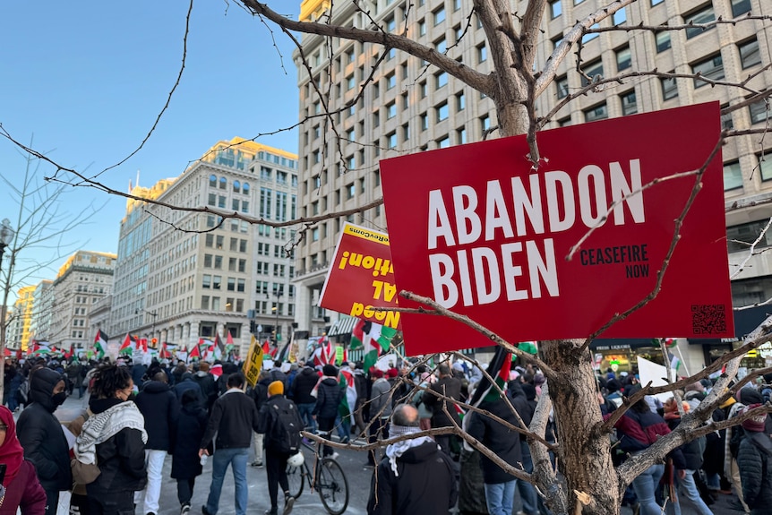 A sign at a March for Gaza reads "abandon Biden" as protestors wave Palestinian flags down a main street