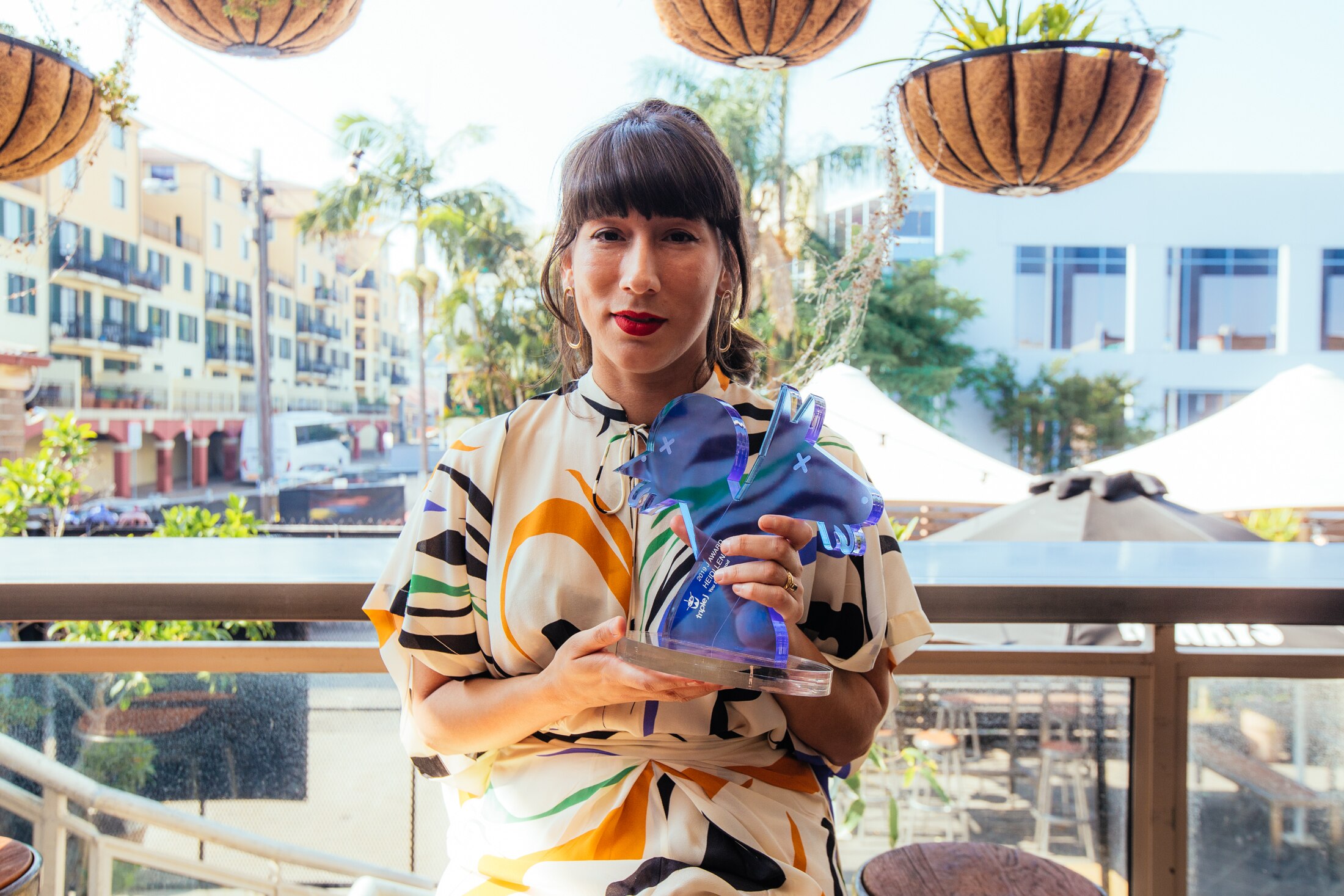 Woman in coloured print dress poses with a transparent blue J Award trophy on the balcony of an inner city venue