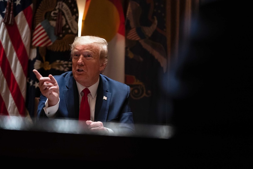 Donald Trump sits behind a desk, pointing to the left