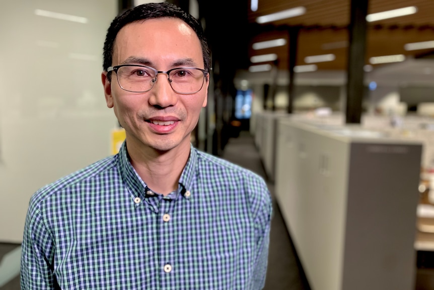 A man wearing glasses stands in an office, smiling at the camera.