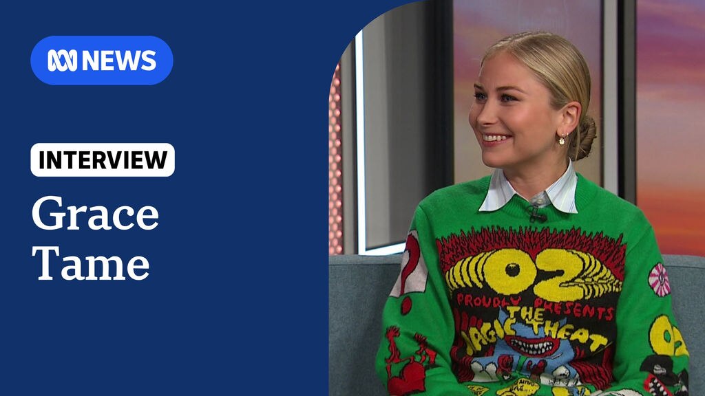 Grace Tame: A woman with blonde hair in a bun and a loud green jumper with cartoon writing on it.