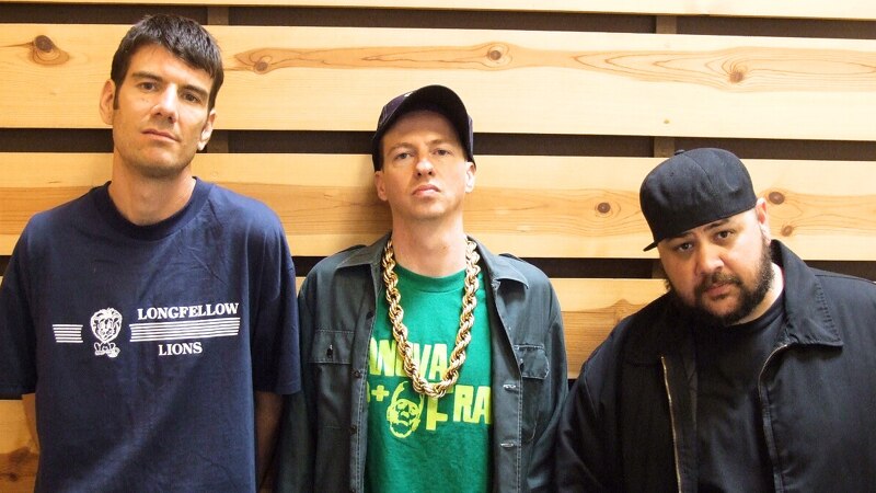 Three members of hip hop group Ugly Duckling stand in front of a wood paneled wall. They all wear t shirts.