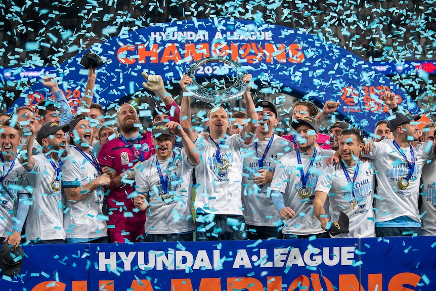 Sydney Fc players celebrate with the A-League trophy as confetti flies around in the air.