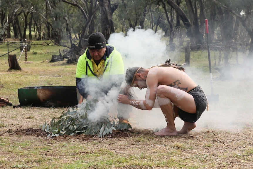 Indigenous people in traditional dress carry out a smoking ceremony in a bush clearing.