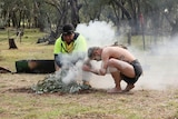 Indigenous people in traditional dress carry out a smoking ceremony in a bush clearing.