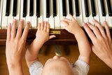An adult and child set of hands play a piano keyboard. The child's hands a placed inside the adults, sitting on a lap.