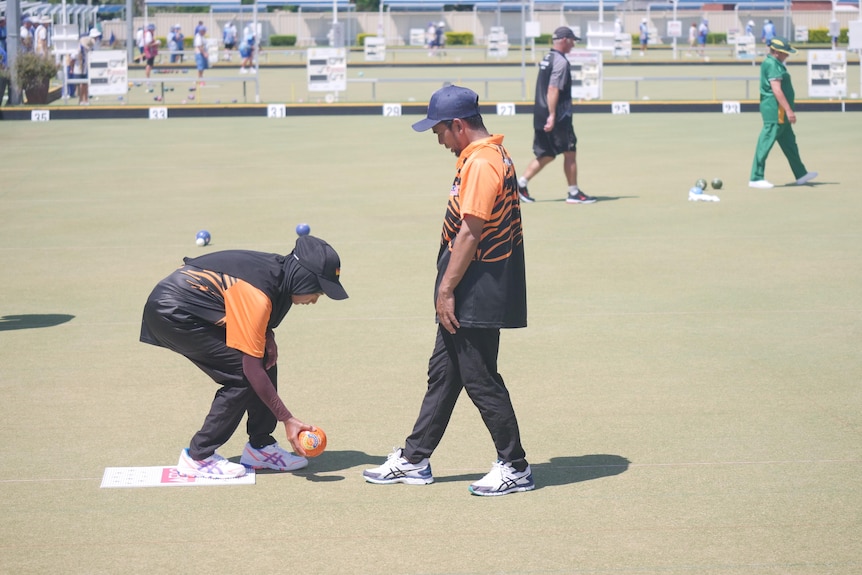 A woman crouches down with a bowling ball in hand while a man stands in front of her with his foot extended.