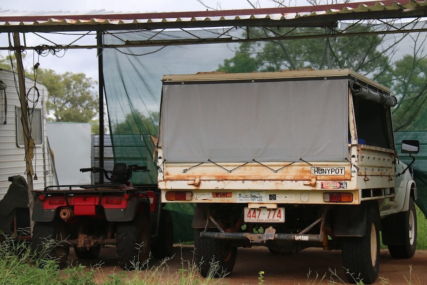 An old ute with a canvas canopy and rusting tray sits next to a quad bike in a makeshift garage.