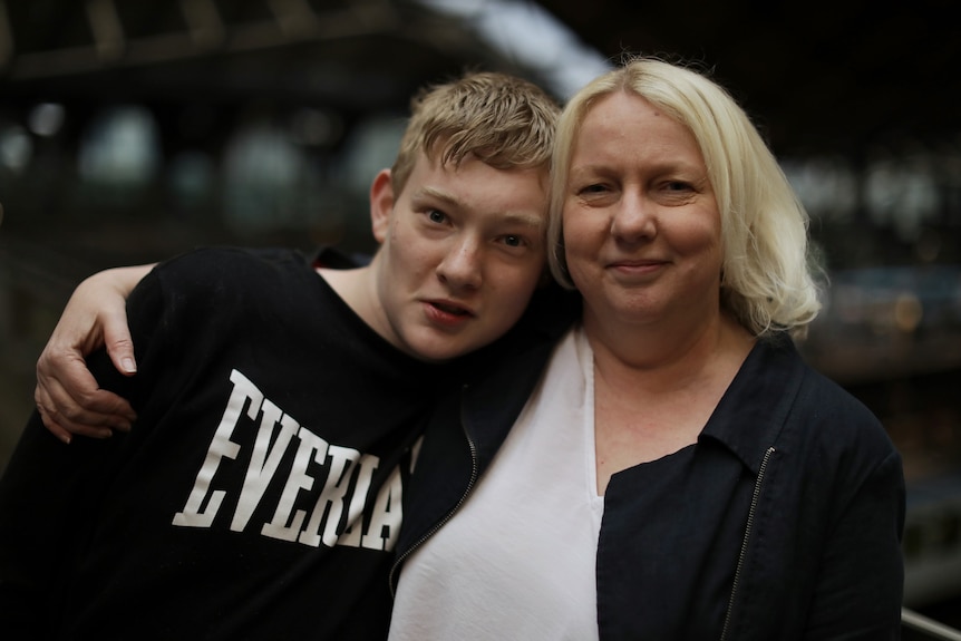 A mother with her arm around her teenage son pulls him close to her. They are both looking at the camera. She is smiling.