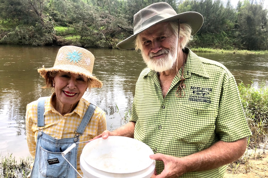 A couple stand in front of a river with a white bucket in their hands.