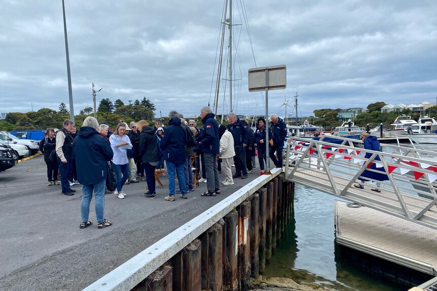 A group of approximately 30 people mill around on a marina wearing jumpers and coats.