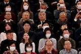 People sitting in a grandstand, some filming with cameras, many wearing masks.