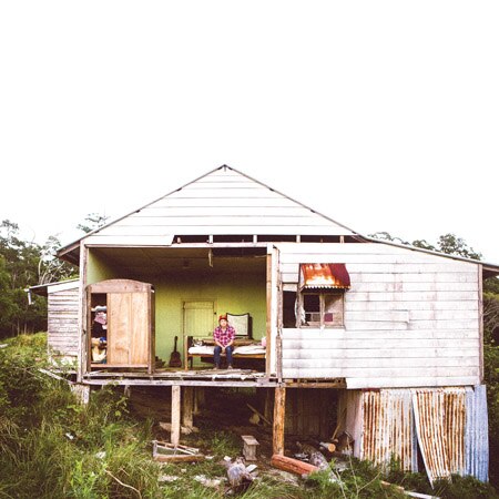 Image of an old house with one wall missing. Darren Hanlon sits on a bed in the house.