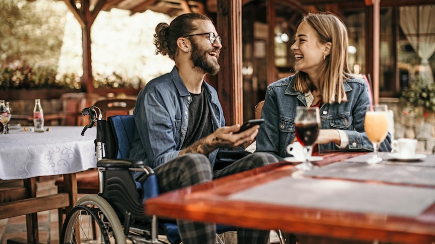 A man in a wheelchair holds a phone and laughs with a woman at a restaurant