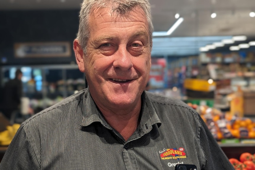 A man has short grey hair, he wears a grey shirt and smiles at the camera, he stands in a vegetable section