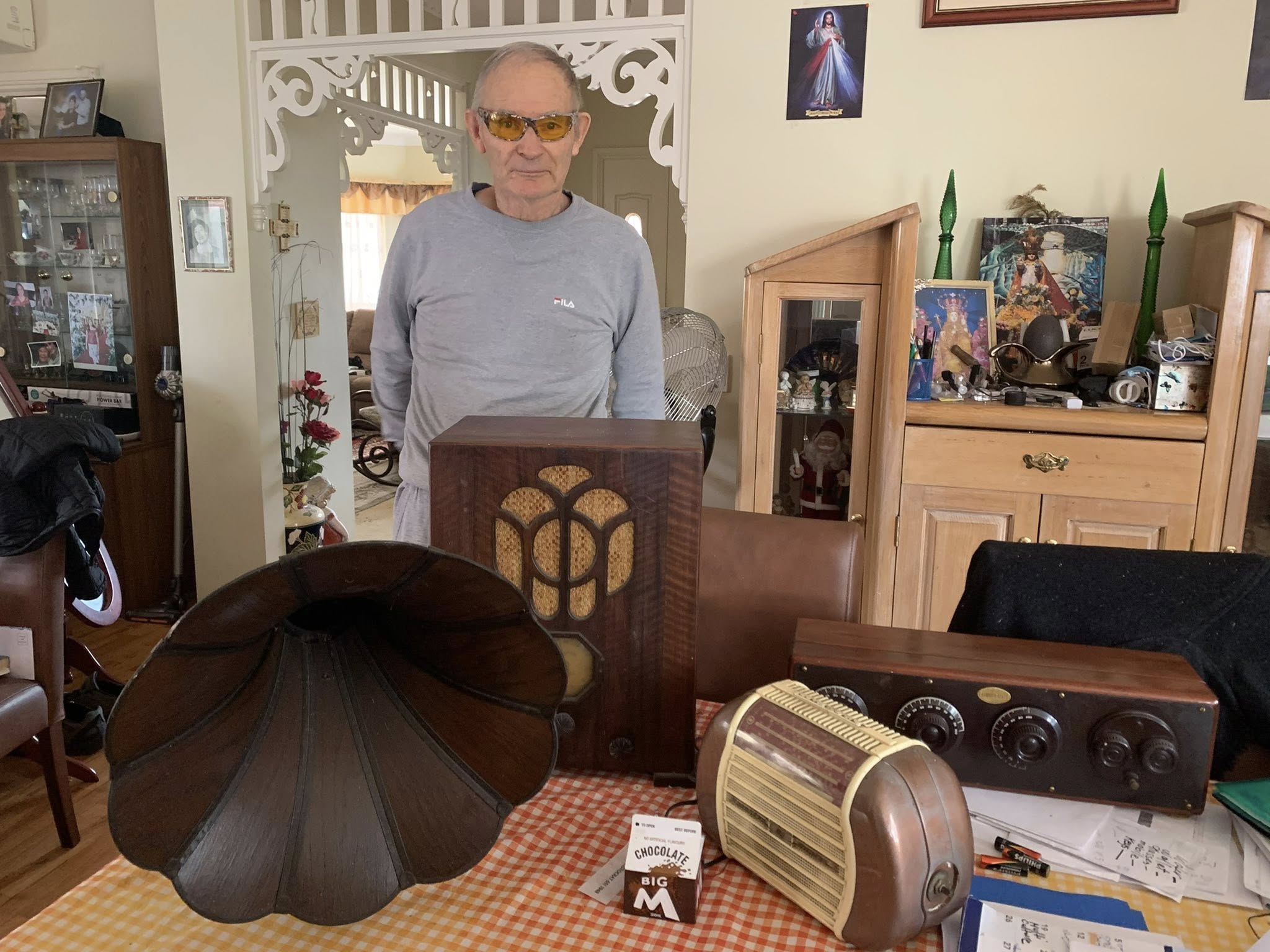 Picture of a man wearing a grey long sleeved shirt and glasses with his collection of vintage radios. 