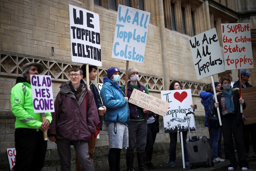 A crowd stands outside a court building, holding placards that read WE TOPPLED COLSTON and GLAD HE'S GONE