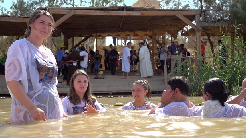 Five people in white shirts wade in a river as other people gather under shelter on the shore.