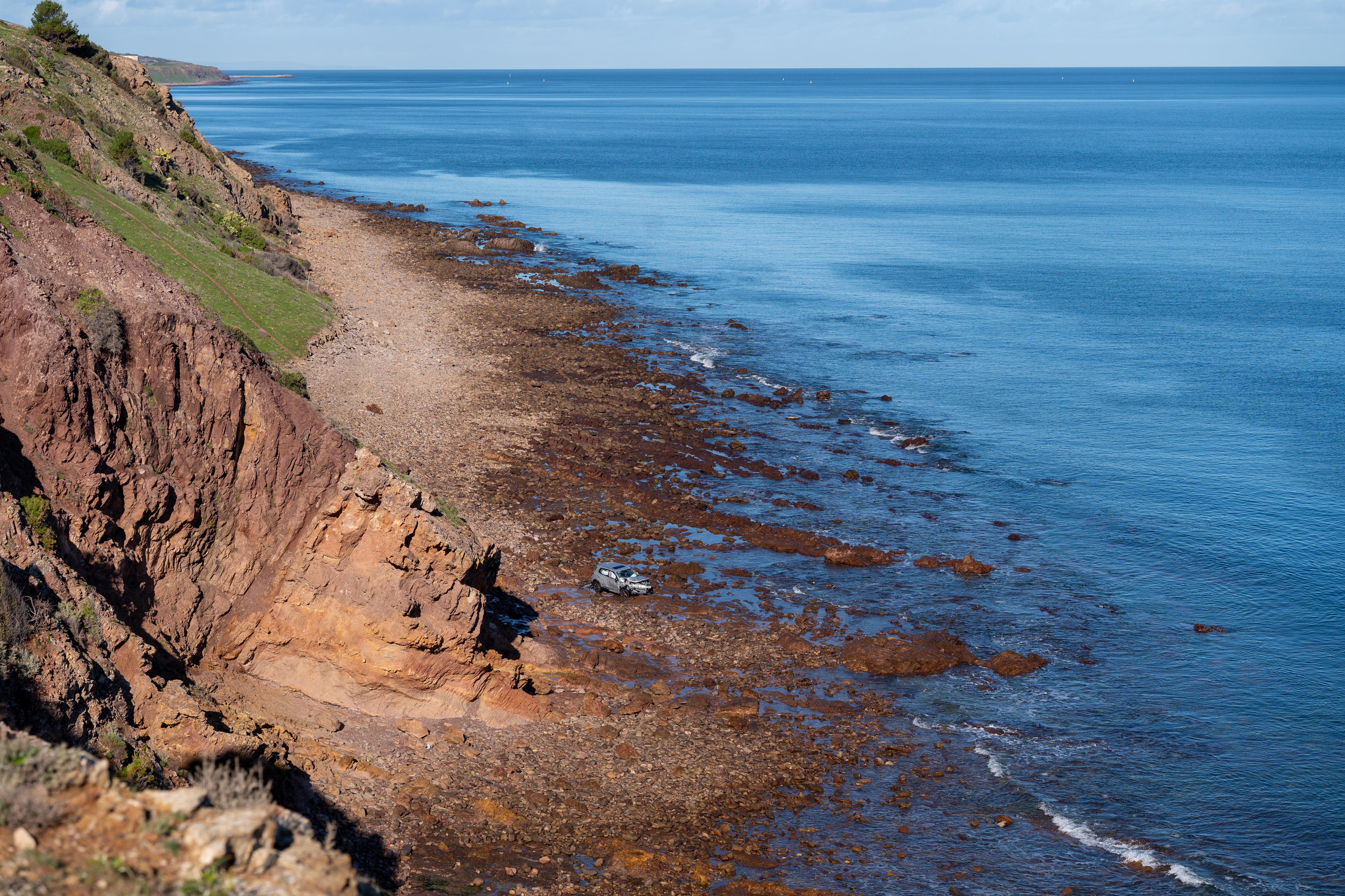 A rocky cliff next to the ocean with rocks on the beach. A car seen as a speck from a distance