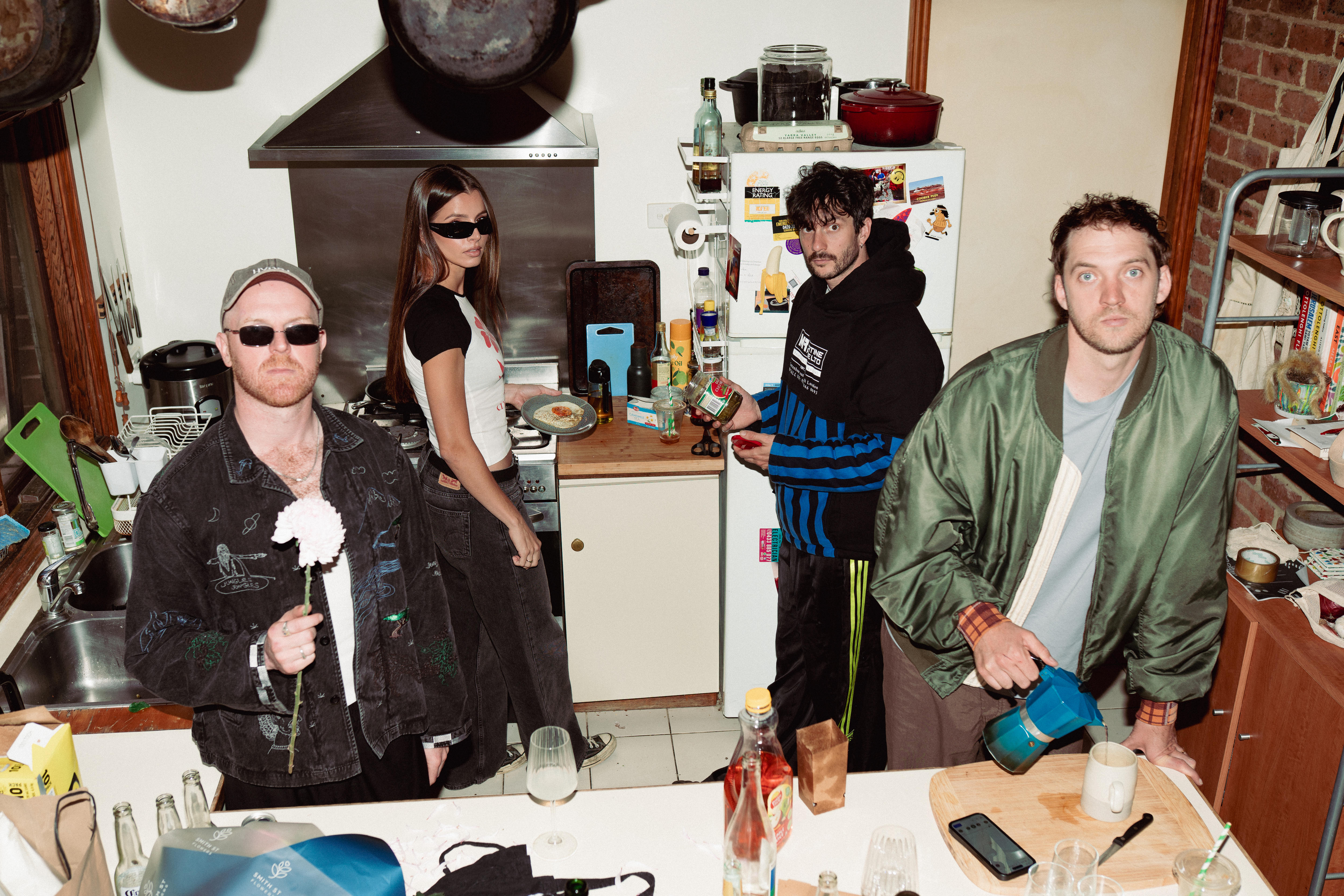 Three men and a woman stand in a busy home kitchen. Twi are wearing sunglasses and they all look up at the camera