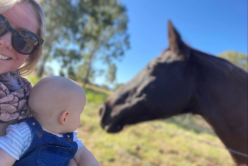 A woman holding a baby, who is looking at a horse 