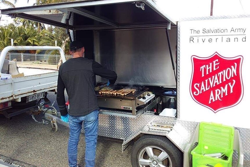 A man dressed in black cooks a line of smoking sausages and onion on a large bbq. He stands next to a Salvation Army sign.
